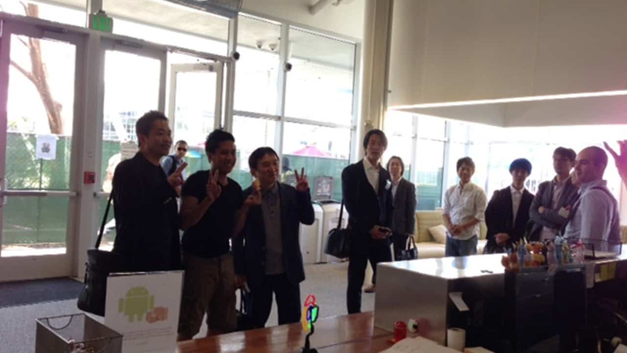 A group of three people posing for a photo inside a building full of windows, making a V sign with their fingers, while others watch from the side
